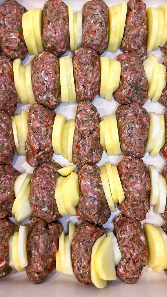 Kafta kebabs in a baking dish with sliced potatoes between