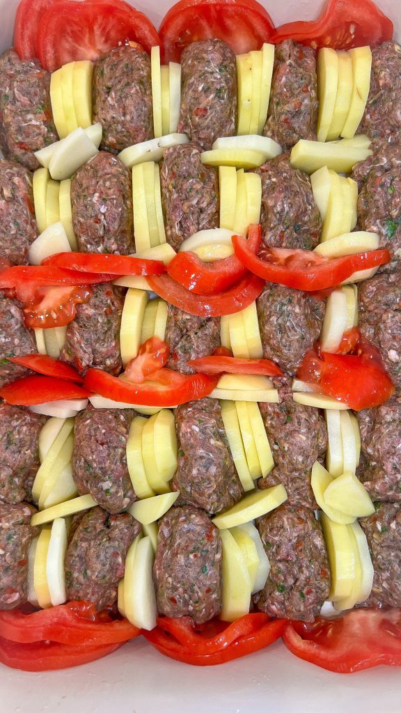 kafta kebabs arranged in a baking dish with sliced potatoes and tomatoes